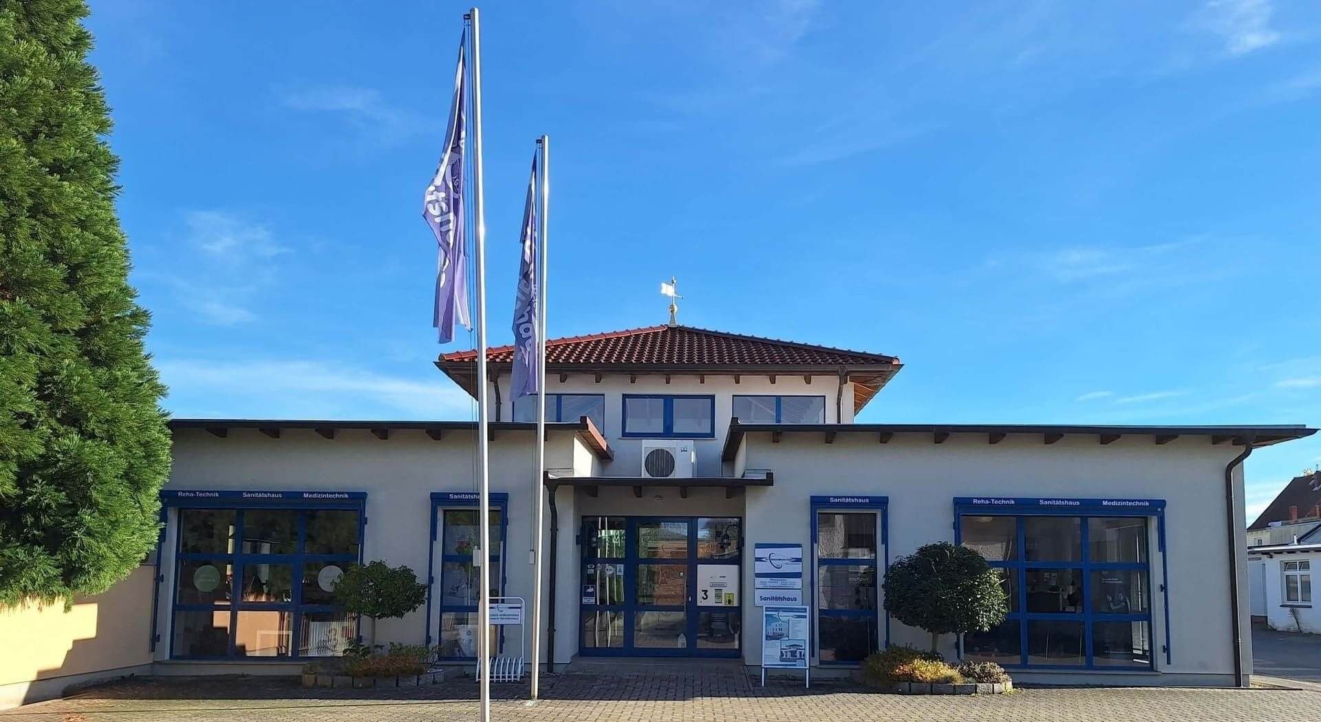 Eingangsbereich eines flachen Gebäudes mit braunem Ziegeldach, drei blauen Türen und Fenstern sowie zwei Fahnenmasten davor